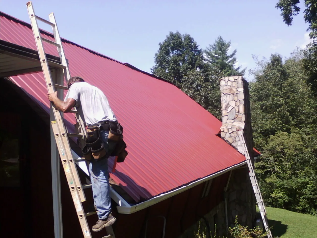 Roofer on a ladder beside a red metal roof and stone chimney — Soffit & Fascia Repair by Roofing Brothers in Carsonville